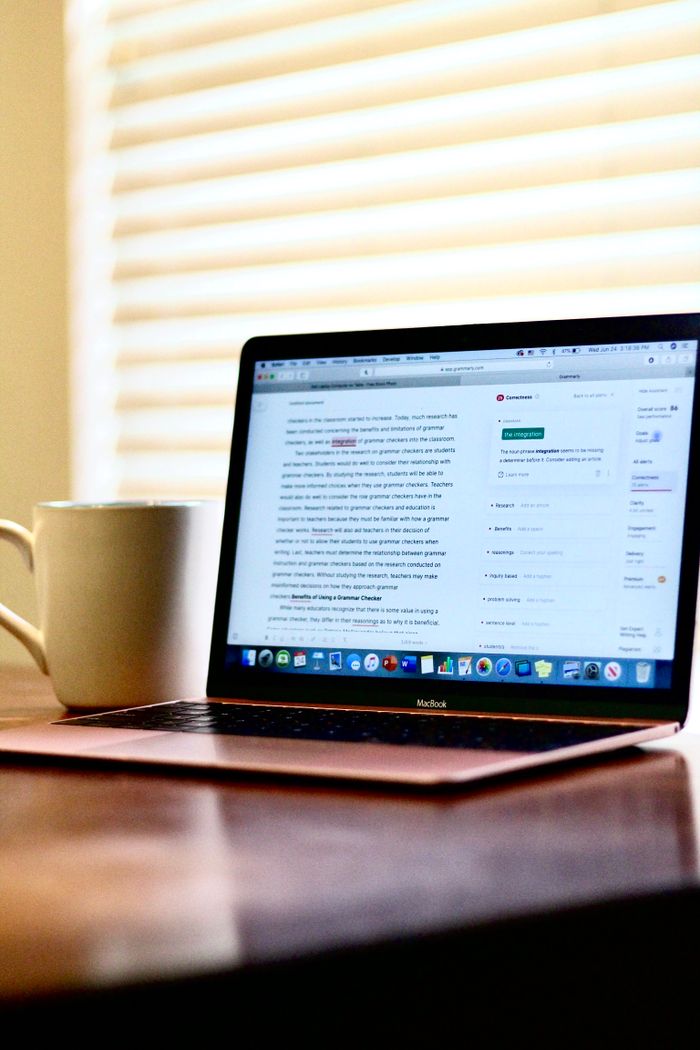 A laptop on a wooden table next to a ceramic mug, illuminated by window blinds.