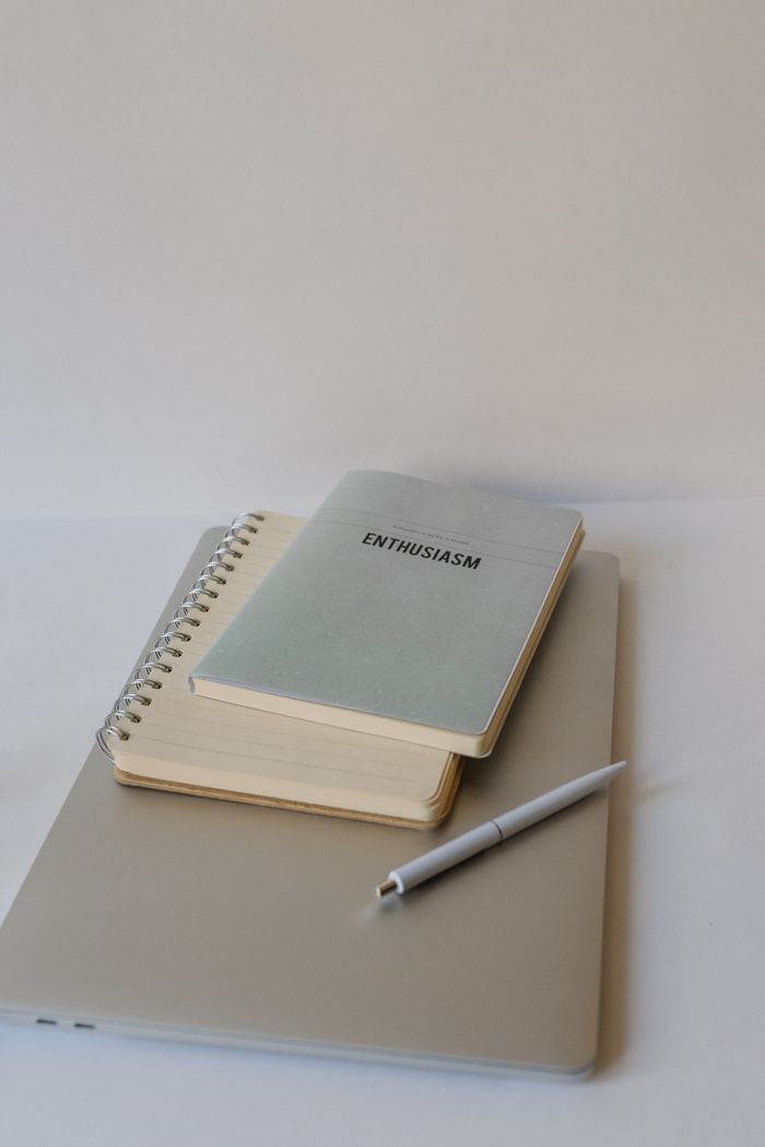 A clean and minimalist office setup with a laptop, notebooks labeled 'enthusiasm', and a pen on a table.